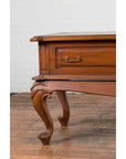 Close-up of a brown wood Dutch Colonial low table with cabriole legs and two drawers, set against a white brick wall.