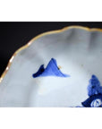 Close-up of blue and white hand painted porcelain bowl with gold rim, scalloped edge, and mountain scene.