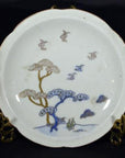 White porcelain plate with hand painted soaring birds and pine trees on brass stand, close-up.