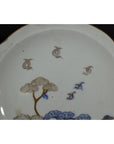 Blue and gold porcelain plate with windswept pines and stylized birds on white background, close-up.