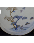 White porcelain plate with hand painted windswept pines and stylized birds, antique Asian design, close-up view.