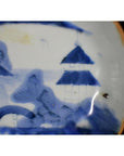 Blue and white porcelain plate with misty peaks, arched bridge, mountains, trees, and dark spot on right side.