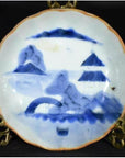 White porcelain plate, hand-painted with "Misty Peaks, Arched Bridge" design, displayed on brass stand.