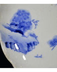 Close-up of blue and white porcelain plate with hand painted mountain landscape and willow trees, scalloped edge.
