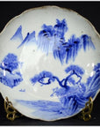 White porcelain plate with hand painted mountain landscape and willow trees, displayed on a stand.