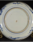 Close-up of underside of white porcelain plate with hand painted pine cone cluster and dense foliage, on brass stand.