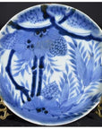 White porcelain plate with Asian design featuring pine cone cluster and dense foliage, on a decorative brass stand.