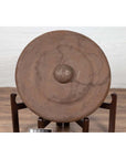 Bronze Burmese temple gong with raised center and red mallet on wooden stand, against a white wall.