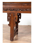 Brown wood Chinese altar console table with overhang top and open fretwork scrolls, on wooden floor by white wall.