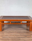Brown Chinese elm coffee table with square legs and rattan inset top, shown on wood floor against white brick wall.
