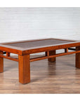 Brown Chinese elm coffee table with rattan inset top and square legs on wooden floor, white brick wall background.