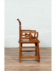 Brown wood hand-carved chair with natural patina on wooden floor against white brick wall indoors.