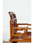 Brown wood chair with carved details on backrest and armrests, placed indoors against a white wall.