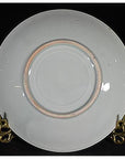 White porcelain plate with gold rim, hand painted and embossed, on a brass stand against a black background.