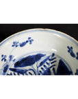Close-up of white porcelain dish with foliate hill motif, rustic brushwork, and chipped rim on black background.