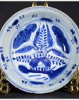 Blue porcelain dish with radial botanical design, displayed on a gold stand against a dark background, close-up.