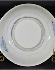 White and blue porcelain plate with seated scholar by river design, on brass stand against dark background, close-up.