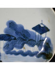 White porcelain plate with painted blue clouded mountain landscape, trees, and a tranquil sloped-roof structure.