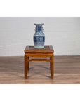 Brown elmwood Ming style side table with horsehoof legs, blue and white ceramic vase on top, against white brick wall.