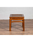 Brown elmwood Ming style side table with horsehoof legs on a wooden floor against a white brick wall.
