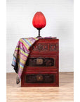 Red and black lacquered antique Chinese chest with carved figures, topped with red lamp and colorful fabric, close-up view.