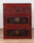 Red and black lacquered antique Chinese three-section chest with carved figures against a white wall.