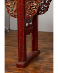 Close-up of red lacquered carved wood table leg with gold accents on a wood floor against a white wall indoors.