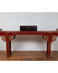 Red lacquered wood console table with carved apron and gold details, displayed indoors against a white wall.