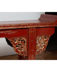 Red lacquered wood console table with carved apron, gold dragon designs, and raised scroll back, indoor setting.
