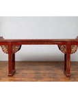 Red lacquered wood console table with carved apron, on wooden floor against plain white wall indoors.