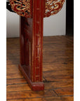 Red lacquered wood console table with carved apron, placed on a worn floor by a white wall in dim lighting indoors.