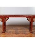 Red lacquered wood console table with carved apron, set on a wooden floor against a plain white wall indoors.