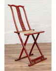 Red lacquered wood folding chair with footrest and fabric, on wooden floor against a white brick wall indoors.