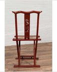 Red lacquered wood folding chair with footrest and fabric, on wooden floor next to white brick wall indoors.