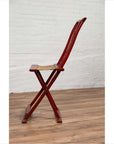 Red lacquered wood folding chair with footrest and fabric, placed indoors against a white brick wall.