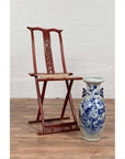 Red lacquered wood folding traveler's chair with footrest next to blue and white vase, indoor setting.