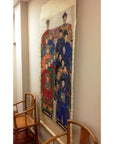 Antique Chinese silk wall hanging in neutral tones displayed between two wooden chairs against a plain wall.