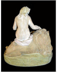 Stone statue of a partially draped figure reclining on a lion, rear view against dark background.