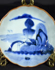 Blue and white hand painted Chinese porcelain bowl with landscape and figures, displayed on gold stand in moody light.