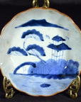 White and multicolor hand painted Chinese porcelain plate with trees, pavilion, mountain, and water on metal stand.