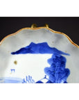 Close-up of blue and white porcelain plate with scalloped edge, hand-painted landscape design, and gold trim.