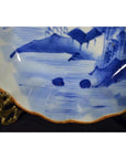 Blue and white porcelain plate with hand-painted landscape, scalloped gold-edged rim, displayed on metal stand.