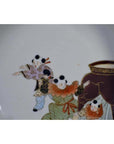 Close-up of an antique hand-painted Japanese Kutani porcelain plate with three figures in traditional dress near a large ornate vase.