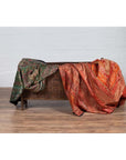 Green and red ornate fabric sarees draped over antique wooden planter on wooden floor, white wall background.