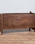 Brown weathered wooden planter with chiseled texture on wood floor against white brick wall indoors.