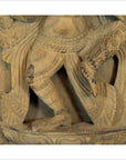 Brown sheesham wood carving close-up showing a figure's bent legs with detailed patterns and traditional clothing.
