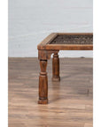Close-up of brown wooden side table with window grate and turned baluster legs on wood floor by white brick wall.