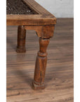 Close-up of brown wooden side table leg with carved details, on wood floor with woven tabletop partly visible.