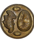 Silver metal Indonesian cake mold with impressions of a curled shrimp, oyster, and two egg shapes, close-up view.