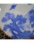 Blue and white ceramic Igezara transferware plate with tree, mountain, and bird design on ornate gold stand.